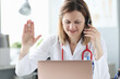 Woman doctor in headphones waving hand at laptop screen