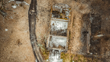 An aerial shot of a rural landscape with a dirt road across a meadow with an abandoned building with a collapsed roof and withered trees. Early spring landscape.