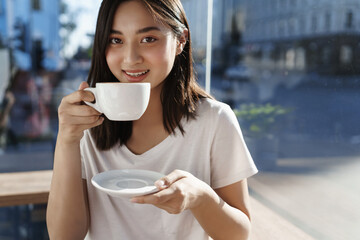 Woman in cafe drinking coffee near window on summer day. Girl sipping cappuccino from cup and smiling at camera, sitting in restaurant