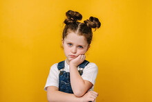 Serious Pensive Little Kid Girl Looking Unhappy At Camera, Tired Upset Preschool Child With Thoughtful Face Holding Hand Under Chin, Melancholic Or Bored, Isolated Over Yellow Studio Background