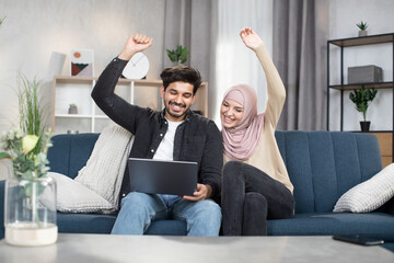 Wall Mural - Cheerful young Muslim couple, holding hands up in victory gesture, celebrating success. Arabian couple using laptop for watching sports match together sitting on comfortable sofa in cozy living room.