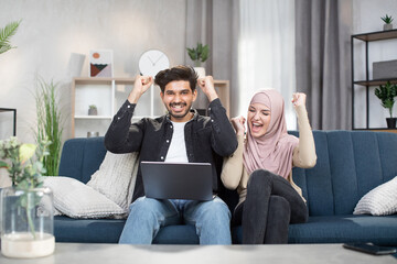 Wall Mural - Joyful Arabian couple of friends, sitting on soaf at home and screaming with raised arms and open mouth, enjoying the winning of favourite sport team. Home, family, sport fans concept.