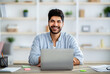 Positive arab male employee smiling at camera while working on laptop at home office
