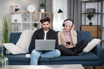 Wall Mural - Handsome bearded Arabian guy sitting on blue sofa with laptop together with his muslim girl in headscarf and earphones, using smartphone and listening to music. Young couple enjoying time at home.