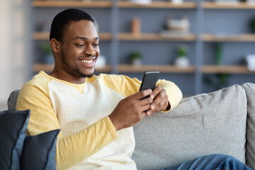 Smiling black guy sitting on couch, using smartphone