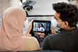 © sofiko14 - Blurred back view of Muslim couple in medical masks, sitting on sofa at home and using tablet pc for online video consultation with their gp family doctor, confident African woman.