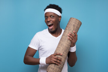 Wall Mural - Happy black man holding yoga mat, smiling at camera over blue wall