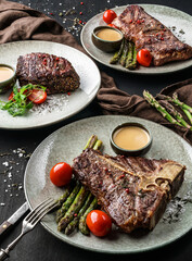 Wall Mural - Grilled tomahawk meat medium rare, rib eye steak, beefsteak on plates with tomatoes, asparagus and berry sauce on dark wooden background. Concept of bbq food, closeup