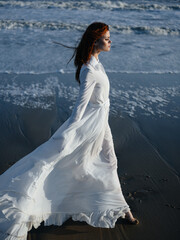 A woman in a white dress walks on the wet sand on the shore of the ocean