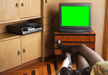 Sexy Girl Sitting In A Vintage Armchair Watches Old TV. An Old Black TV With A Green Screen To Add Video And A VCR Against Old Furniture From The 1980s And 1990s. 1980s And 1990s Apartment.