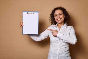 Smiling woman pointing her index finger on white blank paper sheet on clipboard, standing against beige background with copy space