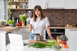 © mdyn - Close up woman in white t-shirt cooking soup with fresh vegetables in kitchen at home. Menu, recipe book banner. Girl reads the recipe in laptop. Caucasian model using internet in loft apartment.