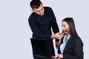 Concentrated male make-up artist makes makeup for a business woman who works while holding a laptop computer. Gray background and side space.