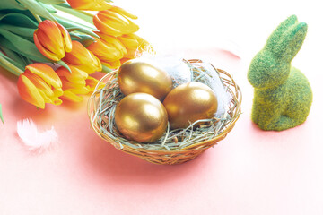 Wall Mural - Basket easter decoration: Golden eggs in basket with spring tulips, white feathers on pastel pink background. Congratulatory easter design.