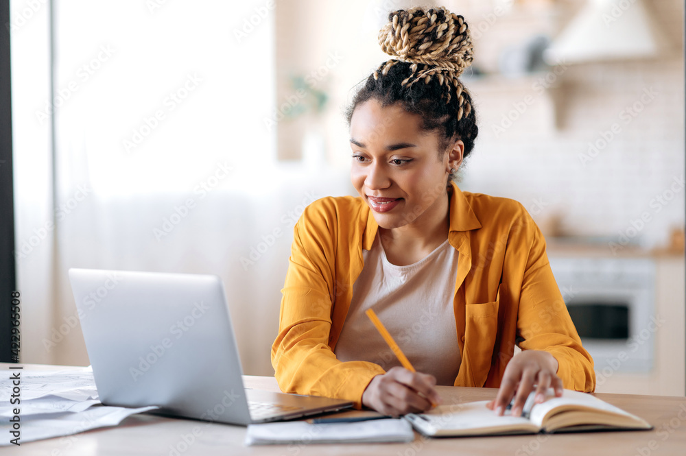 Focused cute stylish african american female student with afro dreadlocks, studying remotely from home, using a laptop, taking notes on notepad during online lesson, e-learning concept, smiling - obrazy, fototapety, plakaty 