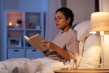 young woman in glasses reading book in bed at home