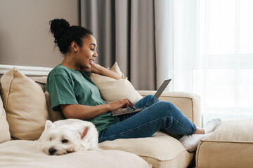 Wall Mural - Smiling black woman using laptop while sitting with her dog on sofa