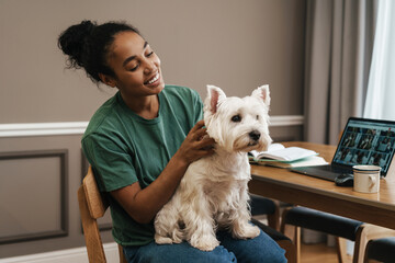 Wall Mural - Smiling black woman stroking her dog while sitting at home kitchen