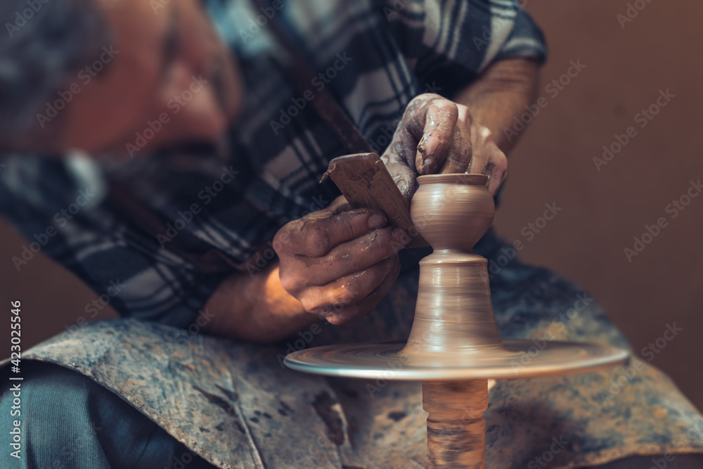 A male artisan in a pottery workshop makes a piece of clay behind a pottery wheel. Traditional pottery craft. - obrazy, fototapety, plakaty 