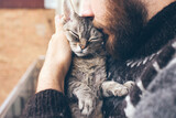 Fototapeta Koty - Close up portrait of a beard man and affectionate Devon Rex cat with closed eyes. Guy is kissing, hugging and cuddling his purring kitty. Feline likes attention and snuggling. Lifestyle photo