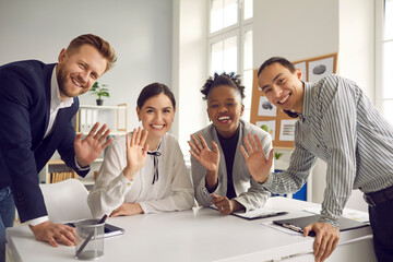 Team of happy young diverse business people sitting and standing around office table, smiling, looking at camera and waving hello, greeting international coworkers in remote work meeting via Internet