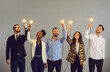© Studio Romantic - Group of happy creative young diverse business professionals holding glowing light bulbs standing on gray studio background. Innovative thinking, finding solution, people developing own idea concept