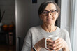 © Vadim Pastuh - Charming elderly woman wearing stylish eyeglasses stands with cup of morning coffee looks at the camera and smiles, friendly female with mug of hot drink spends time at home