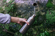 Crop Anonymous Hiker Filling Metal Bottle With Fresh Cold Natural Spring Water From Source With Pipe In Mountainous Forest