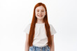 Cute little kid with long red hair smiling and looking happy at camera, standing over white background. Girl child express joy and positive emotions