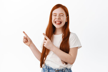 Wall Mural - Funny ginger little girl points upper left corner aside, smiles and shows tongue with pleased childish face expression. Redhead kid showing advertisement with happy emotion, white background