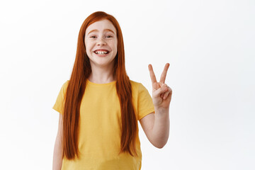 Wall Mural - Beautiful redhead girl with freckles smiling broadly, showing two fingers peace sign and looking happy, standing in yellow t-shirt against white background