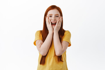 Wall Mural - Surprised ginger girl with freckles checking out something awesome, stare with amazement and joy at surprise gift, standing happy against white background