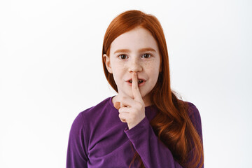 Wall Mural - Shh this is secret. Cute redhead little girl, child shushing, show keep quiet taboo gesture, finger on lips, hushing, standing over white background