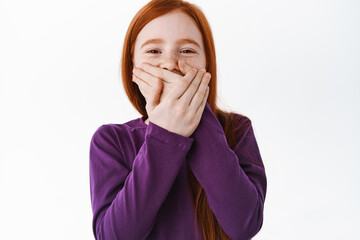 Wall Mural - Portrait of beautiful red head girl with freckles laughing, covering her mouth with hand and chuckle over funny thing, joking, standing over white background