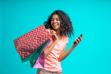 Wall Mural - Attractive african american young woman with perfect smile is posing with a pile of paper shopping bags and mobile phone in hands