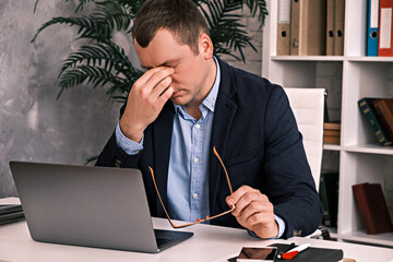 Eye fatigue, poor eyesight, office work. A young man takes off his glasses and rubs his eyes with his hand while sitting at a table with a laptop in a suit. Slow motion