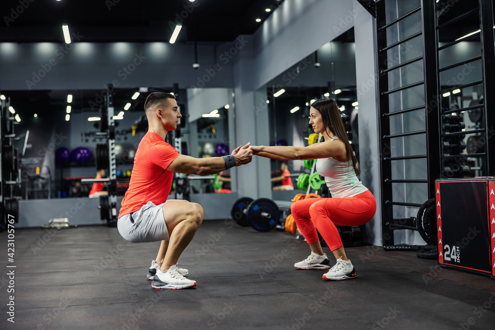Two athletes, a beautiful woman and a handsome man are in a squatting position in the gym or fitness center. They look each other in the eyes and hold hands. Fitness lover, eye contact - obrazy, fototapety, plakaty 