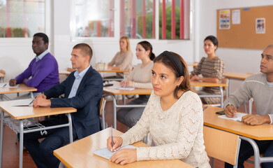 Portrait of focused young adult female sitting at desk studying in classroom with colleagues