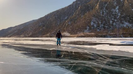 Wall Mural - Cool man in blue pants, a helmet, sunglasses, ice skates and ski poles is rolling on the ice of frozen Lake Baikal. A beautiful winter landscape with clear, smooth blue transparent ice and deep cracks