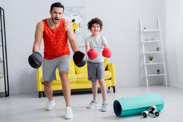 Wall Mural - Excited muslim father in punch mitts standing near son and sport equipment at home