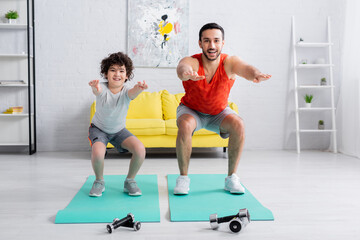 Wall Mural - Smiling arabian son and father doing squats on fitness mats near dumbbells