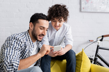 Wall Mural - joyful muslim man pointing at cellphone near smiling son