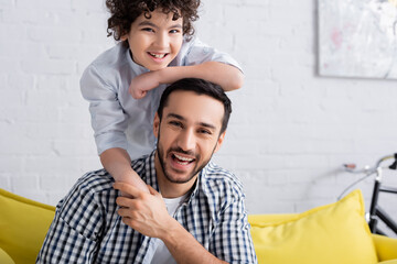 laughing muslim man holding hand of smiling son and looking at camera