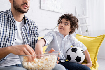 Wall Mural - smiling arabian kid holding soccer ball and taking popcorn near father watching football match