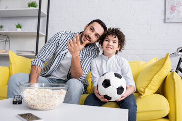 smiling muslim man pointing with hand near son while watching football championship at home