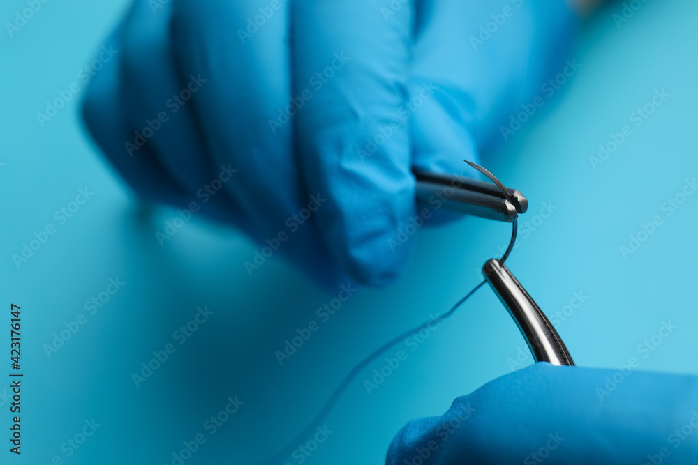 Professional surgeon holding forceps with suture thread on light blue background, closeup. Medical equipment - obrazy, fototapety, plakaty 