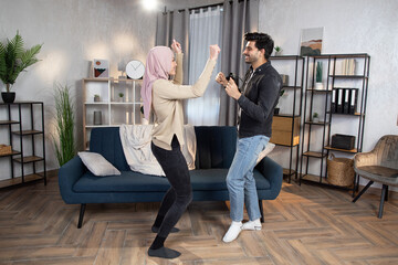 Wall Mural - Having fun and nice time at home concept. Bearded young Arabian man dances with his pretty wife in a hijab, spending free evening time together at home living room.