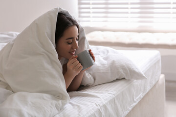 Sticker - Beautiful young woman covered with warm blanket enjoying hot drink on bed at home. Space for text