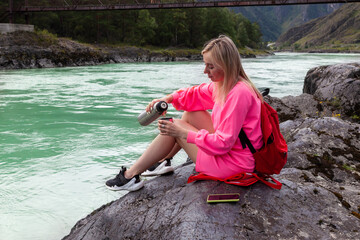 a young beautiful blonde girl in a pink suit with shorts sits on a large stone near the river in the Altai mountains and pours hot tea from an iron thermos into a mug while relaxing