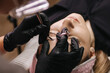 eyebrow microblading. A master in a transparent mask on his face and black gloves holds a handpiece with a shading needle over the eyebrow of the model.